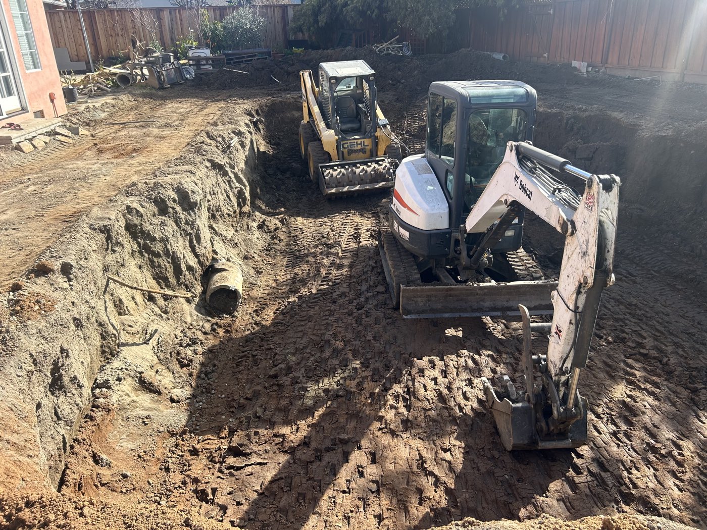 Skid steer and mini excavator excavating a pool cavity with a drain pipe staged nearby