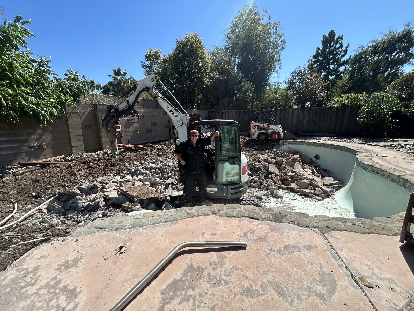 Terry standing with his mini excavator on a tight, fenced backyard pool-removal job site
